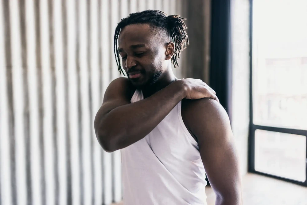 Black man holding his shoulder with a slight expression of discomfort, symbolizing oxidative stress and the wellness benefits of a glutathione push therapy.